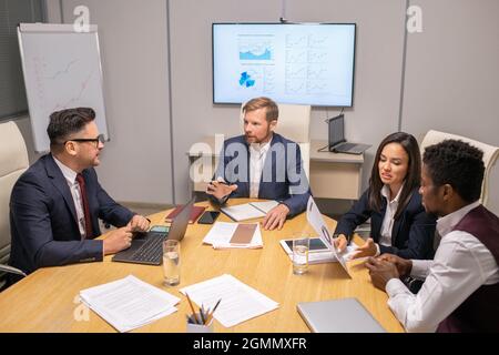 Two businessmen consulting about speech points while two intercultural colleagues discussing papers by table in boardroom Stock Photo