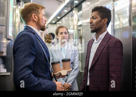 Two intercultural businessmen and young businesswoman standing in moving elevator Stock Photo