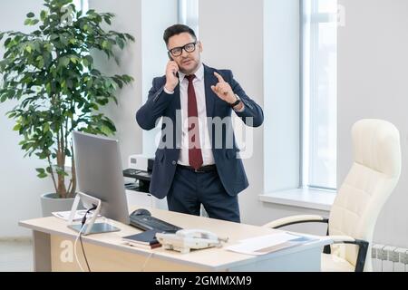 Confident employer in elegant suit discussing terms of deal on telephone while standing by his workplace Stock Photo