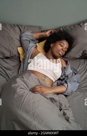 Young pretty African woman sleeping under blanket in bed with grey pillows