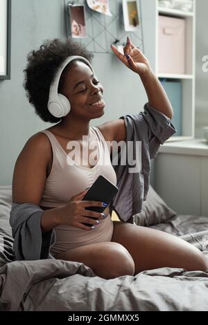 Young excited female with headphones enjoying her favorite music while sitting in bed Stock Photo