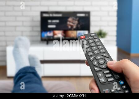 Man watching TV, lying on sofa, legs on table. Person holding remote control in living room Stock Photo