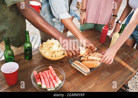 Group of young intercultural friends taking fresh grilled hotdogs while standing by wooden table Stock Photo
