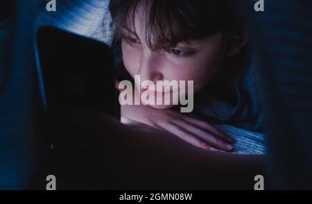 Happy teen girl using smartphone, hiding under blanket at nigh, social networks cocnept. Stock Photo