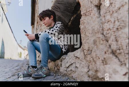 Low angle view of preteen girl with smartphone sitting outdoors in town. Stock Photo