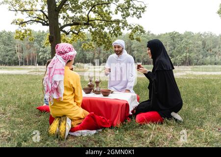 Cheerful arabian family in traditional clothes holding tea in park Stock Photo
