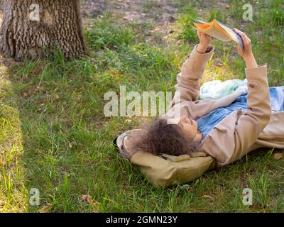 Photo of a mature adult woman reading a book in the park. She lies on the gras with a book and take a break from constantly working at the computer. D Stock Photo