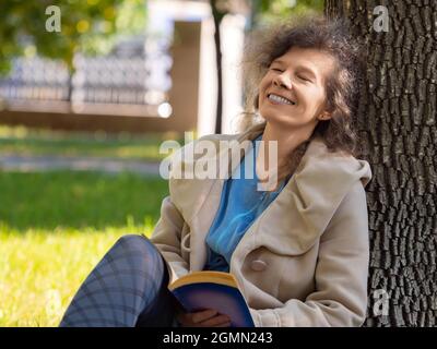 Photo of a middle-aged woman with a book in the park. She sitting on the grass in city park with a book and smiling with closed eyes. Concept of rest Stock Photo
