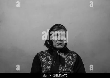 Defocus portrait of 60s russian grandmother senior old woman seating indoors. Old women in shawl. Sad person. White dirty background. Black and white. Stock Photo