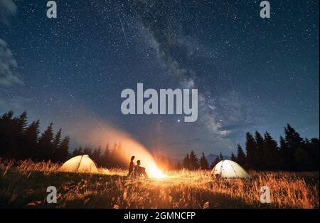 Magnificent view of night starry sky over mountain valley with illuminated camp tents and hikers near campfire. Young woman and man travelers sitting near bonfire. Concept of night camping and galaxy. Stock Photo