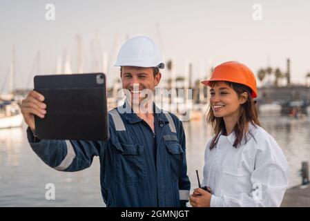 Positive professional male and female engineers in protective helmets having video call via tablet while working in sea port together Stock Photo