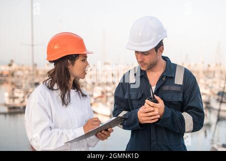Professional female inspector filling checklist on clipboard while communicating with male employee with VHV radio hands during inspection of sea port Stock Photo