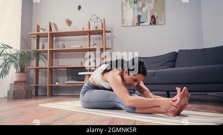 Fit sportswoman touching legs while stretching on fitness mat in living room Stock Photo