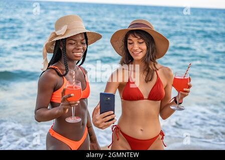 Charming cheerful young multiracial females in bright colorful bikinis and hats sharing photos on social networks while chilling on beach with drinks during summer holidays Stock Photo
