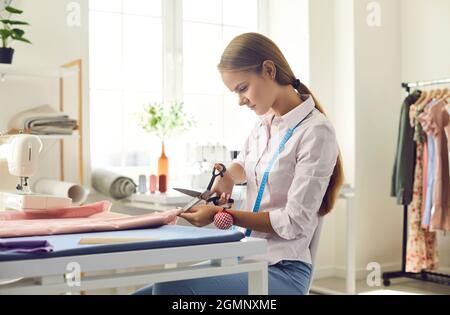 Focused seamstress with scissors makes marking cuts on fabric while working in clothing studio. Stock Photo