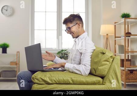 Happy young man sitting on couch with laptop computer and having online work meeting Stock Photo