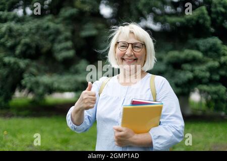 Smiling blonde teacher woman lady 40s 50s years old wearing eyeglasses standing hold notepads looking camera outdoors Stock Photo