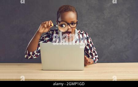 Dark-skinned woman with a surprised expression looks at the laptop screen through a magnifying glass Stock Photo