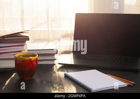 Opened laptop, books and cup of coffee on old wooden desk in morning sunlight. Cosy home workspace in morning Stock Photo
