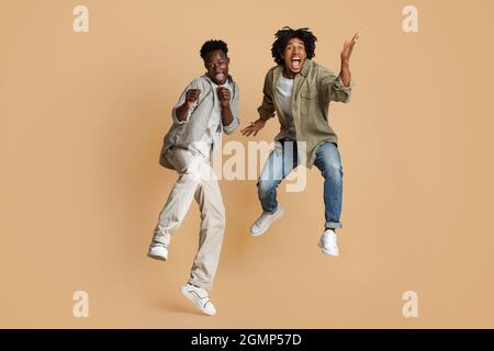 Crazy Mood. Two Overjoyed Black Guys Jumping Over Beige Studio Background Stock Photo