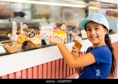 A little girl is standing near a shop window with an assortment of ice-cream and sweets. The child chooses sweets in the display case. City lifestyle Stock Photo