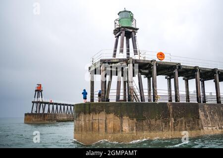 Whitby harbour with anonymous men fishing on the edge of the pier wall during rough seas and high tide. Colourful red and green lighthouse towers. Stock Photo