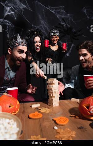 excited african american woman playing wooden tower game with multiethnic friends in halloween costumes on black Stock Photo