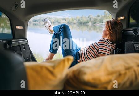 Woman dressed in warm calm colors clothes and jeans lying in the cozy car trunk and enjoying the mountain lake view. Warm early autumn auto traveling Stock Photo