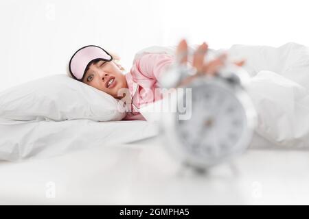 Funny sad tired millennial caucasian blonde lady in pink pajama and sleep mask reaches to alarm clock, lies on bed Stock Photo