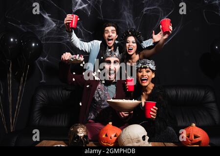 thrilled multicultural friends in halloween costumes near popcorn, carved pumpkins and skulls on black Stock Photo
