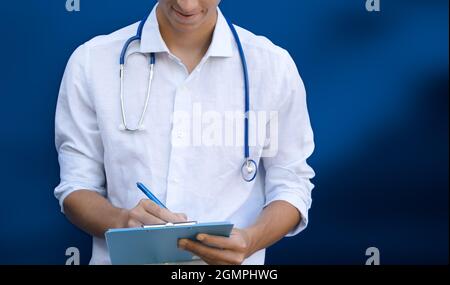 Young doctor writes on clipboard. Blue background with copy space Stock Photo