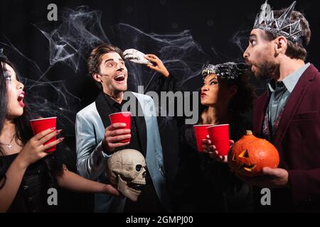 african american woman in wolf mask scaring man with toy hand near multiethnic friends on black Stock Photo