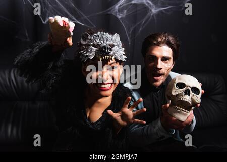cheerful african american woman in wolf mask showing scary gesture near man holding skull on black Stock Photo