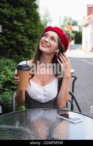 happy young woman in red beret, earphones and eyeglasses holding paper cup near smartphone with blank screen Stock Photo
