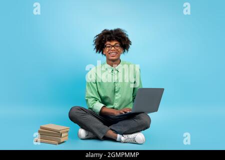 Excited black teenager with laptop and books nearby, sitting on floor on blue studio background, free space Stock Photo