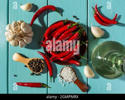 Directly above view of raw red hot chili pepper pods, sea salt and whole black peppercorns in wooden spoons, garlic head and olive oil glass jar. Stock Photo