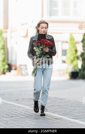 full length of pretty woman in leather jacket holding red roses and walking on urban street in europe Stock Photo
