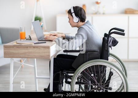 Side view of handicapped teen boy in wheelchair wearing headphones, having video chat with teacher or classmate at home Stock Photo