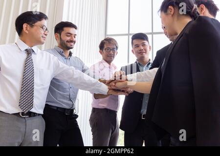 From below of multiethnic business men and women stacking hands together while standing in office in back lit Stock Photo