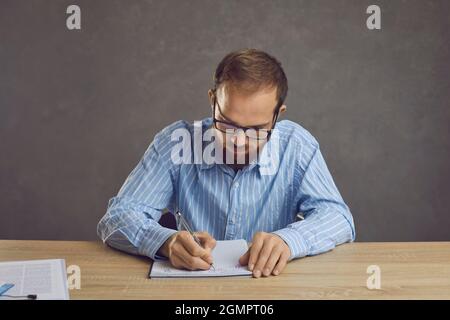 Serious young man in glasses sitting at a table and taking notes in his notebook Stock Photo