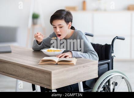 Focused disabled teenage boy in wheelchair eating cereal for breakfast and reading book at home. Domestic hobbies Stock Photo