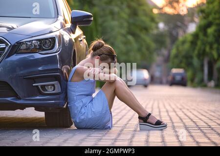 Young woman driver sitting beside her broken car waiting for help. Vehicle problems concept. Stock Photo