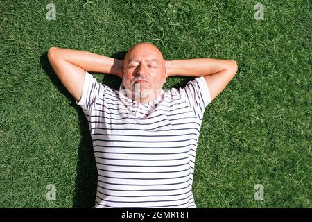 Retired man lying on his back on grass with hands on head with grass background. Stock Photo