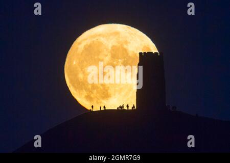 Glastonbury, Somerset, UK.  20th September 2021.  UK Weather.  The full Harvest Moon rises up into the clear night sky from behind St Michael’s Tower on Glastonbury Tor in Somerset silhouetting the people on the summit.  Picture Credit: Graham Hunt/Alamy Live News Stock Photo