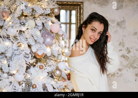 a young attractive brunette woman, wearing a white sweater pulled down over one shoulder near the Christmas tree. The girl smiles at the camera. The mood of the holiday. Stock Photo