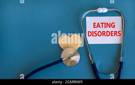 Eating disorders symbol. White card with words Eating disorders, beautiful blue background, wooden heart and stethoscope. Medical and eating disorders Stock Photo