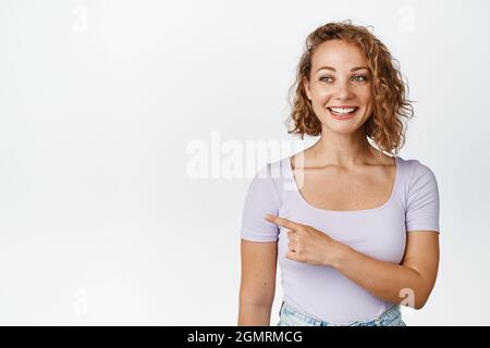 Smiling blond woman with curly short hair, pointing finger and looking left at banner with happy face expression, showing sale advertisement, white Stock Photo