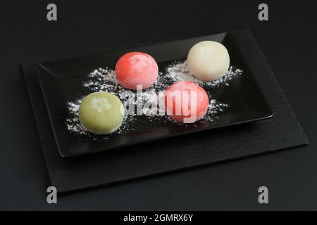 Four types of Japanese dessert mochi - pomegranate with honey, green matcha tea, strawberry, coconut on a black plate on a black table. Closeup. Stock Photo