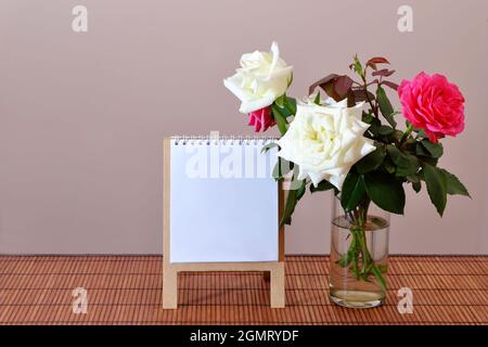 A bouquet of white and red roses in a glass on a bamboo tablecloth with a white sheet of paper on a stand. Mockup. Selective focus. Stock Photo