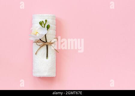 White towel roll tied with rope with sprig of orchid flower on pink paper background. Copy space Template for lettering text or your design. Stock Photo